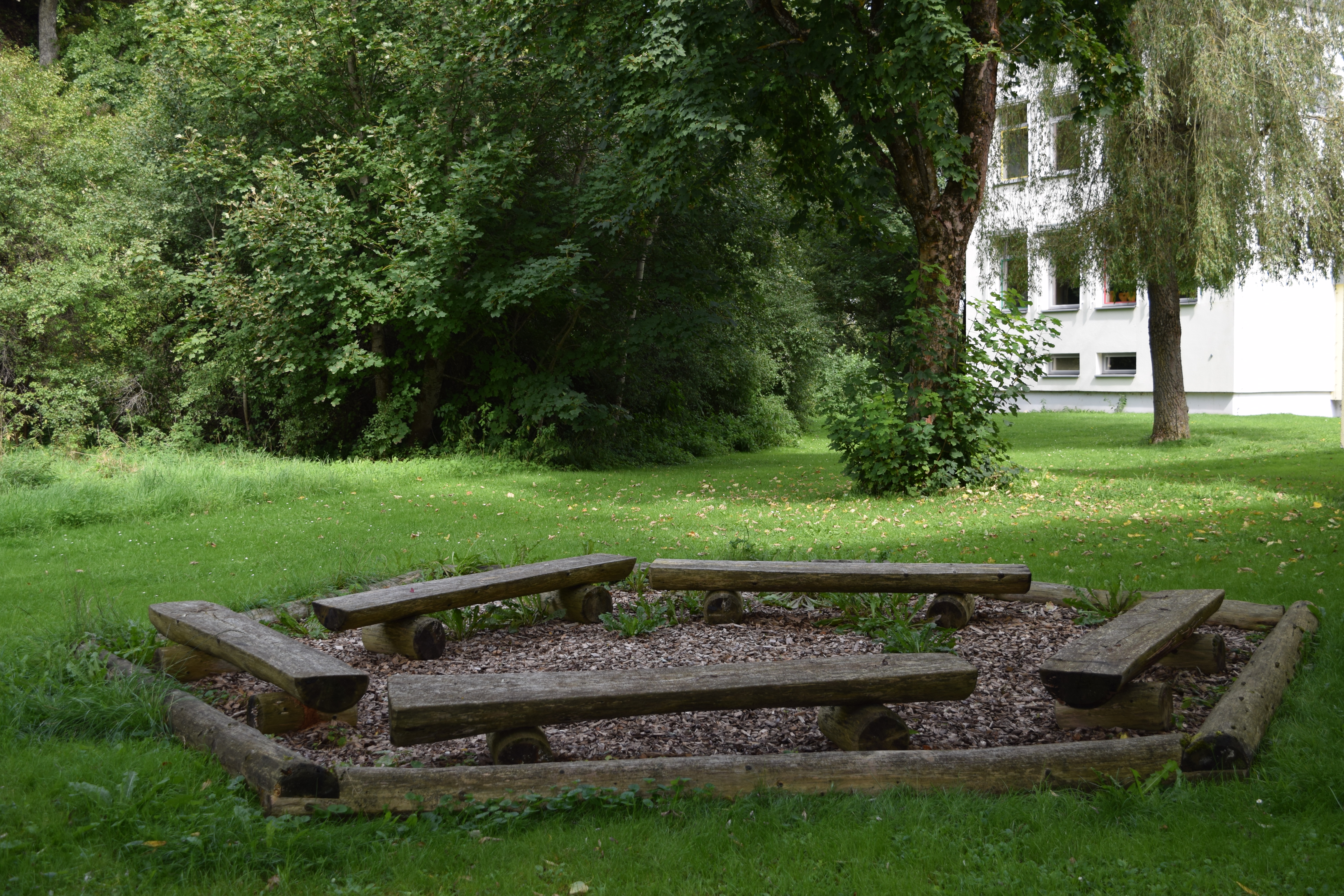 Auf diesem Bild ist ein Garten mit Holzbänken, die im Fünfeck angeordnet sind, zu sehen. Ein Klassenzimmer im Grünen. 